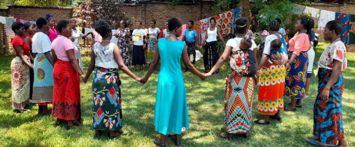 Caregivers in Training stand in a circle, practicing the games they will use to teach children later on. They wear colourful prints and some have babies on their backs.