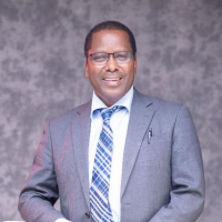 Dezie is a black man with thin square glasses and short, tight curly hair. In this photo he wears a grey suit with a white shirt and blue striped tie. He is smiling brightly at the camera.